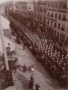 Photo en noir et blanc de militaires défilant dans une rue d'un centre-ville avec des spectateurs les observant