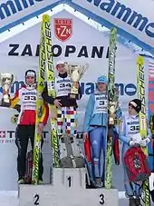 Quatre jeunes femmes en tenue de sport et dossard numéroté de compétition tenant des skis de saut verticalement d'une main, dont trois sur un podium chacune sur une marche, tenant chacune une coupe en métal doré d'environ 50 centimètres, et une à leur gauche, sur fond pavoisé de marques de sponsors et marqué « ZAKOPANE »