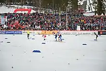 Vue de loin et de profil de fondeuses disputant l'arrivée d'une course, devant une tribune.