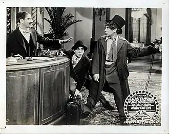 Photographie en noir et blanc représentant trois personnages dont l'un, en tenue débraillée et coiffé d'un haut de forme brandit un modèle ancien de combiné téléphonique