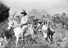 Photo en noir et blanc montrant un homme sur un cheval blanc, à sa suite des hommes marchant avec des sacs et des lances