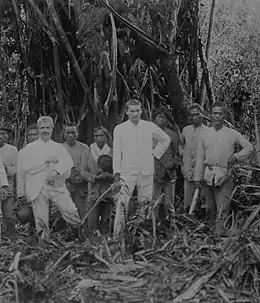 Photo en noir et blanc représentant un groupe d'hommes de face en bordure d'une clairière, dos à la forêt ; deux hommes blancs habillés en blanc posent devant sept Indonésiens de plus petite taille.
