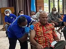 Un médecin, penché sur l'épaule d'un homme assis, injecte un vaccin