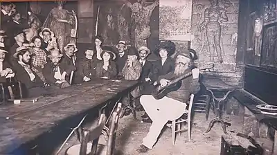 Photo sépia d'une une longue table, un groupe de gens écoutant un homme barbu à la guitare