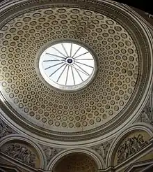 L’intérieur de la coupole à caissons du Cabinet d’Histoire Naturelle. La verrière est au centre, en bas deux tympans sculptés des portes avec entre eux une des voûtes en cul-de-four. Éclairage zénithal par la verrière. Décors à dominante blanche, saufs les caissons et le fond des tympans en stuc jaune.