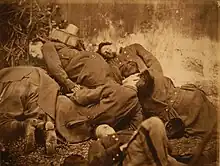 Photographie sépia mise en scène, montrant six hommes en tenue militaire allongés les uns sur les autres, les yeux clos, devant un mur lépreux.