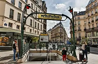 Photographie d'une bouche de métro avec trois femmes au premier plan, une image publicitaire pour les Folies Bergères sous le nom de la station et une devanture à l'enseigne d'une célèbre chaîne de Hamburgers à l'arrière-plan