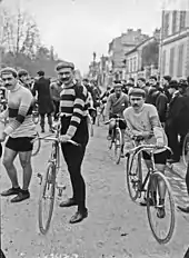 Photographie en noir et blanc montrant plusieurs cyclistes se tenant debout à côté de leurs vélos avant le départ d'une course.