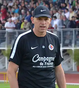 Photographie d'un homme avec un t-shirt noir et une casquette noire. En arrière-plan, des spectateurs en tribune