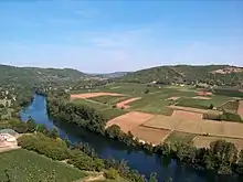 Photo couleur. Prise du haut d'une falaise, elle montre un méandre de la rivière (ou cingle) qui enserre des prairies et des vignes dans une presqu'île.