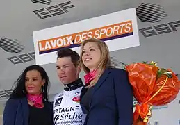 De trois-quarts, trois personnes sourient. Un coureur au maillot blanc tient un bouquet orange dans sa main gauche.