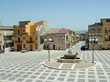 Photographie couleur d’une place de village avec une fontaine au centre.