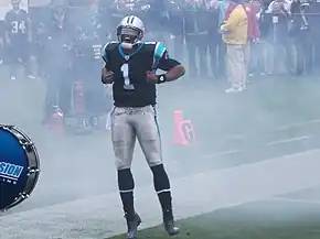 Cam Newton en tenue de football américain, poings fermés, les écartant depuis le centre de sa poitrine.