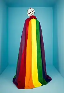 Une longue chute de tissu parcourue verticalement de larges bandes d'arc-en-ciel posée sur un mannequin en plastique vu de dos.