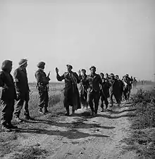 Photo en noir et blanc de militaires marchant sur un sentier avec les mains en l'air et de d'autres militaires se tenant debout à côté du sentier les regardant