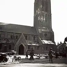 Le même jour, un soldat canadien faisant la circulation devant l'église de Bernières-sur-Mer. Sont visibles sur l'édifice des impacts d'obus et des dégâts sur la toiture.
