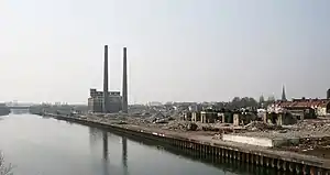 Photographie en couleur avec, au premier plan, un canal, et au second plan, à gauche un bâtiment industriel et deux cheminées d'usine et à droite un terrain vague jonché de gravats et de vestiges de constructions