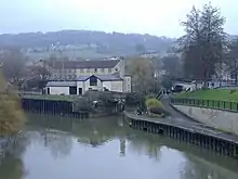 Une rivière coule lentement à partir du milieu à gauche vers le bas à droite, avec l'entrée d'un canal vers le coin supérieur droit. Le canal passe immédiatement sous un petit pont, et à 100 mètres plus loin, sous un autre pont. Une colline avec des maisons forment l'arrière-plan.