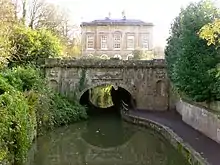 Un bâtiment en pierre jaune. Sur la gauche des arbres tombent dans l'eau.