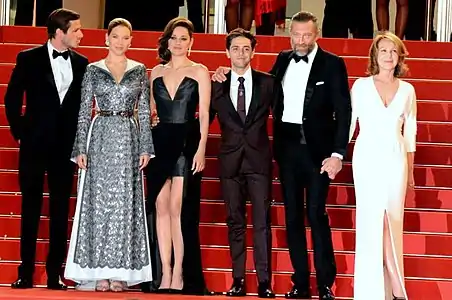 Six personnes élégamment vêtues se tiennent côte à côte sur les marches d'un tapis rouge.