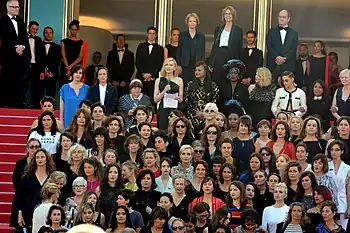 Photographie de plusieurs dizaines de femmes en tenue de gala alignées sur les marches du palais des festivals à Cannes.