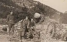 Quatre militaires assis devant un gros canon à large roues, dans un paysage montagneux.