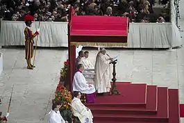Le trône papal et le pape François derrière son pupitre sous un dais aux couleurs pourpres et aux parements dorés