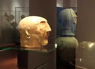 Tête stylisée en céramique avec un crâne allongé, une boucle d'oreille en or et un menton assez long, des trous et des fissures sont visibles.