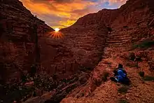 Reliefs arides. Deux personnes regardent le soleil disparaître derrière la montagne, assis sur un promontoir au dessus d'une vallée dont on ne voit pas le fond.