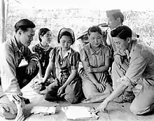 Photographie montrant quatre femmes assises en tailleur, discutant en train de regarder des documents disposés au sol. Elles sont entourées par deux hommes en uniformes.