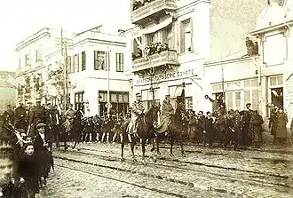 Photographie sépia : des troupes à cheval dans une rue.