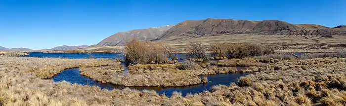 Un cours d'eau bordé de plantes sèches serpente dans une large vallée bordée de montagnes dans un grand soleil.