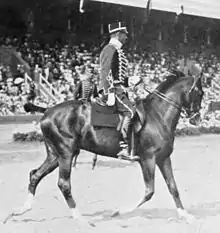 Photo en noir et blanc représentant un couple au trot sur une carrière; le cavalier est en uniforme et tient les rênes d'une seule main.