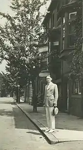 Photographie en noir et blanc d'un homme en costume clair avec cravatte, tenant un chapeau clair, debout dans une rue avec des arbres.