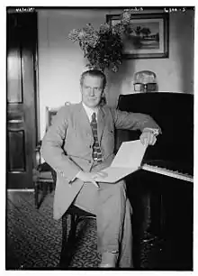 Portrait en noir et blanc d'un homme assis en costume, un livre sur ses genoux, appuyé contre un piano