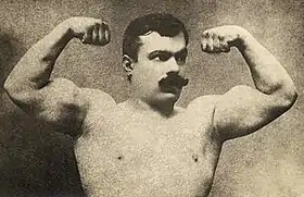 Un homme à moustaches torse-nu montre ses biceps.