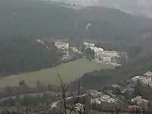 En plongée : quelques bâtiments modernes proches d'un château moderne, à la jonction d'un pré et de forêts, au fond d'une vallée. Les pavillons résidentiels voisins occupent le quart inférieur droit de la photo.