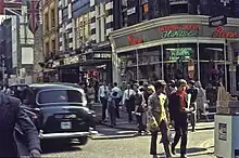Colour photo of a busy city intersection with two young white males walking across in the foreground