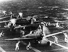 Photo en noir et blanc d'avions sur le pont d'envol d'un porte-avions, moteur tournant.