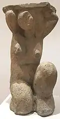 Statue de pierre de couleur grise, d'une femme nue accroupie ayant les bras levés au-dessus de la tête