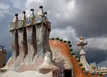 En quatre plans : groupe de cheminées polychromes, toit de la Casa Batlló, croix gaudienne, ciel d'orage.