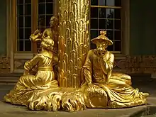 Une sculpture dorée représentant une colonne autour de laquelle évoluent trois personnages, une femme, un homme et une personne portant un chapeau de paille.