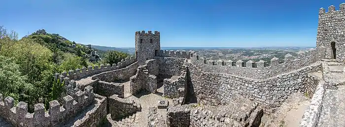 Une muraille crénelée ondule sur d'un périmètre emplit de petits murs en ruines séparant des pièces. Au dehors des arbres, au loin une plaine avec une ville.