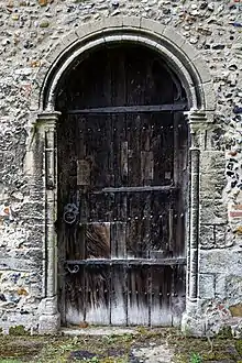 Photographie couleur d'une porte de bois forgé, encastrée dans un mur de pierre et dont le seuil donne sur un sol conquis par de petites herbes.