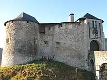 Vue de l’accès d’un château fort, avec une tour ronde à gauche et une tour carrée surplombant l’entrée avec pont-levis.