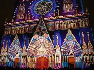 Vue en couleurs d’une façade de cathédrale, prise de nuit et illuminée de multiples couleurs.