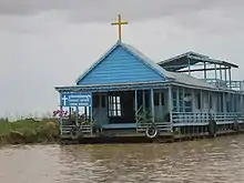 Le bâtiment est de couleur bleu avec une croix au sommet du toit au-dessus de l'entrée. À gauche, un panneau avec l'inscription église catholique de Chong Khneas en khmer et en anglais.