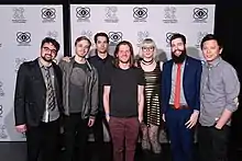Sept personnes se tiennent devant un panneau de photographie et sourient à la caméra.