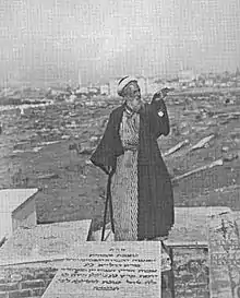 Un rabbin âgé en tenue traditionnelle, on distingue des pierres tombales avec des inscriptions en hébreu devant lui. D'autres tombes du cimetière en arrière-plan.