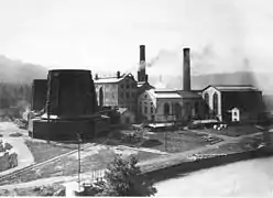 Photo noir et blanc montrant de hauts bâtiments industriels accompagnés de deux tours de refroidissement métalliques et de deux cheminées d'usine.